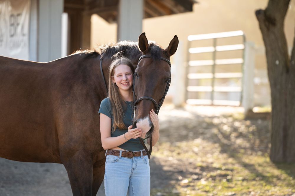 Person wearing jeans and standing next to a brown horse while sunlight streams through barn slats.