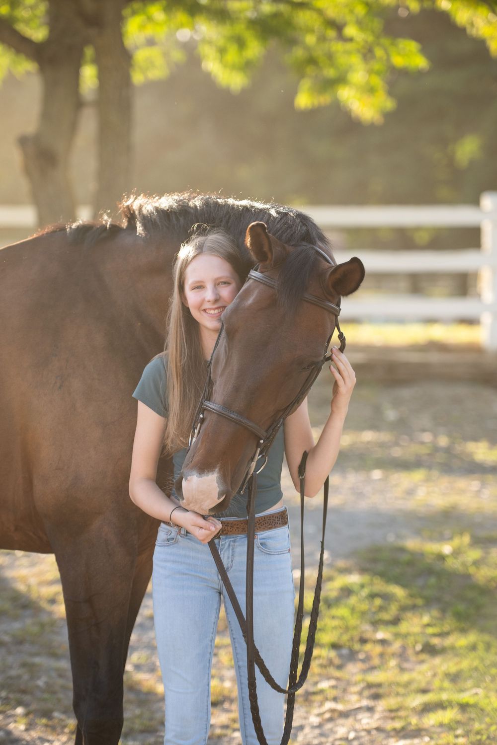 A horse and rider share a tender moment outdoors during golden hour with sunlit trees in the background.