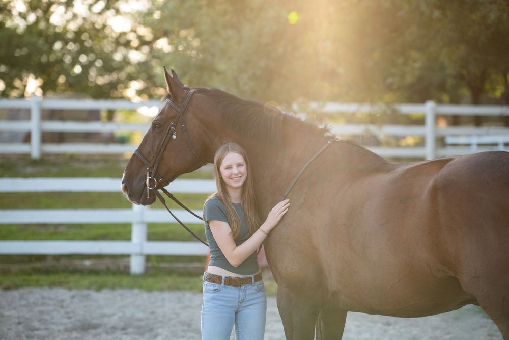 Woman leading brown horse along white fence during sunset with sunflare in background.