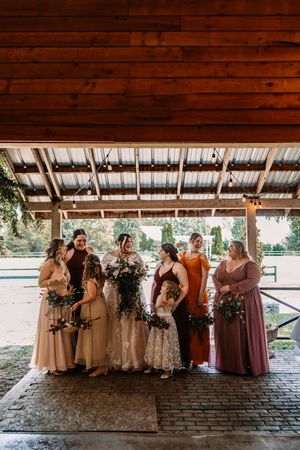 Bride with bridesmaids in mixed earth-tone dresses holding bouquets under rustic wooden pavilion.