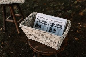 White wicker basket holding custom wedding newspaper programs on wooden stool outdoors.