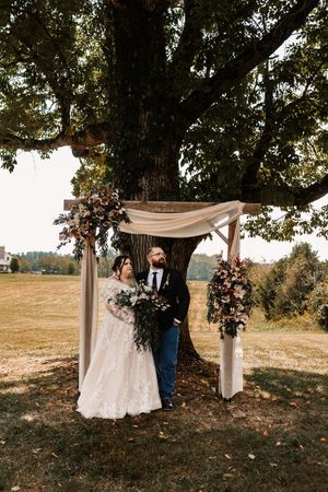 Bride and groom standing together under floral wooden arch beside large oak tree.