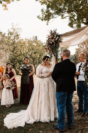 Bride in lace gown raising bouquet during outdoor wedding ceremony under large oak tree.