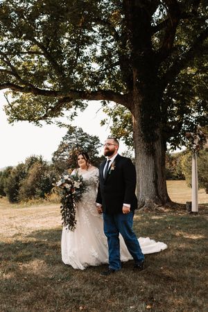 Bride and groom walking hand in hand beneath large oak tree after outdoor wedding ceremony.
