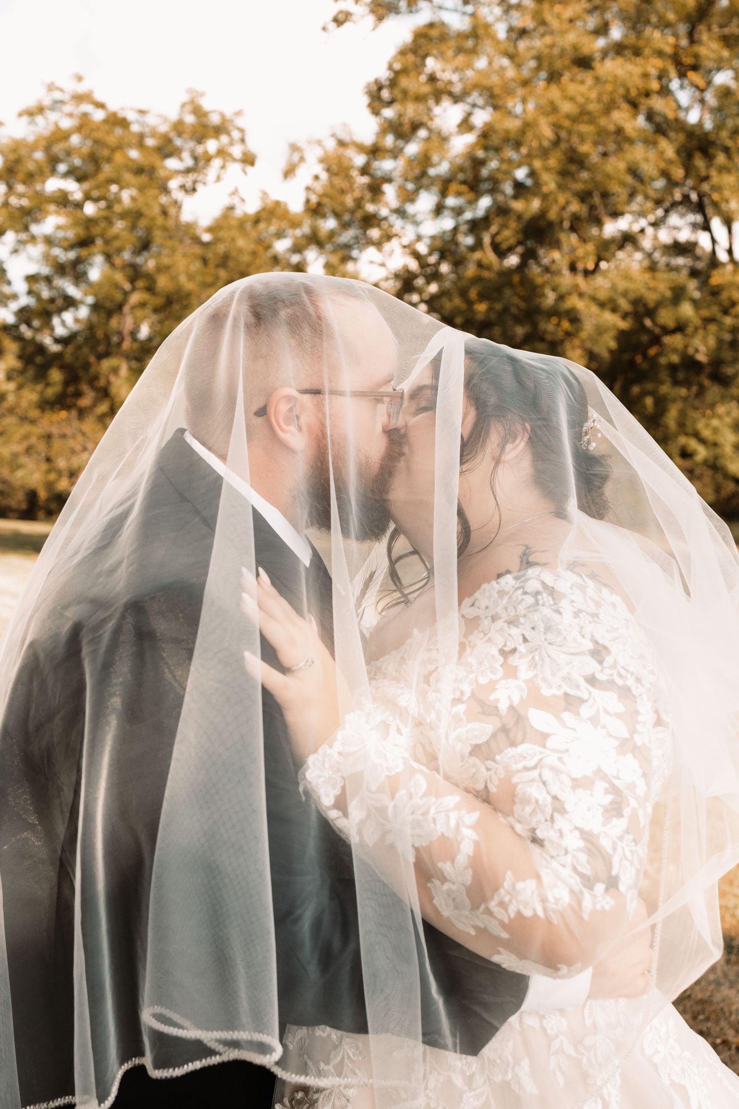 Bride and groom sharing a kiss under a flowing veil outdoors among autumn trees.
