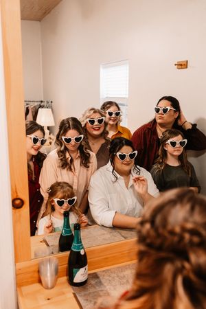 Bride and bridesmaids wearing heart-shaped sunglasses celebrating with champagne in mirror selfie.