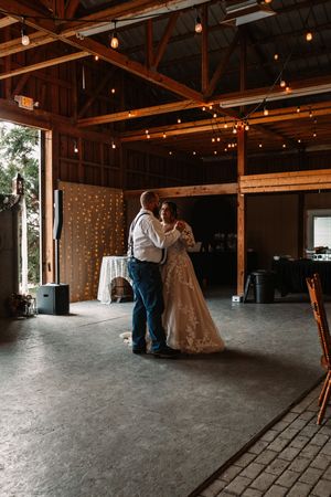 Bride and groom sharing first dance in rustic barn venue with warm string lights overhead.