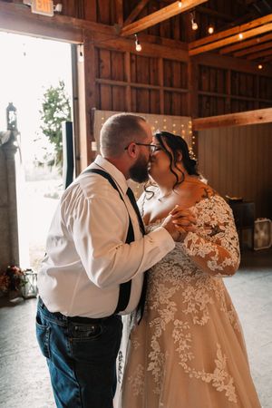 Bride and groom share a kiss during their first dance inside a rustic barn wedding venue with string lights.