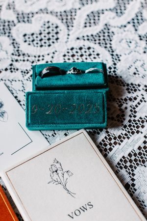 Teal velvet ring box with wedding date 9-20-2023 holding two rings beside vows booklet.