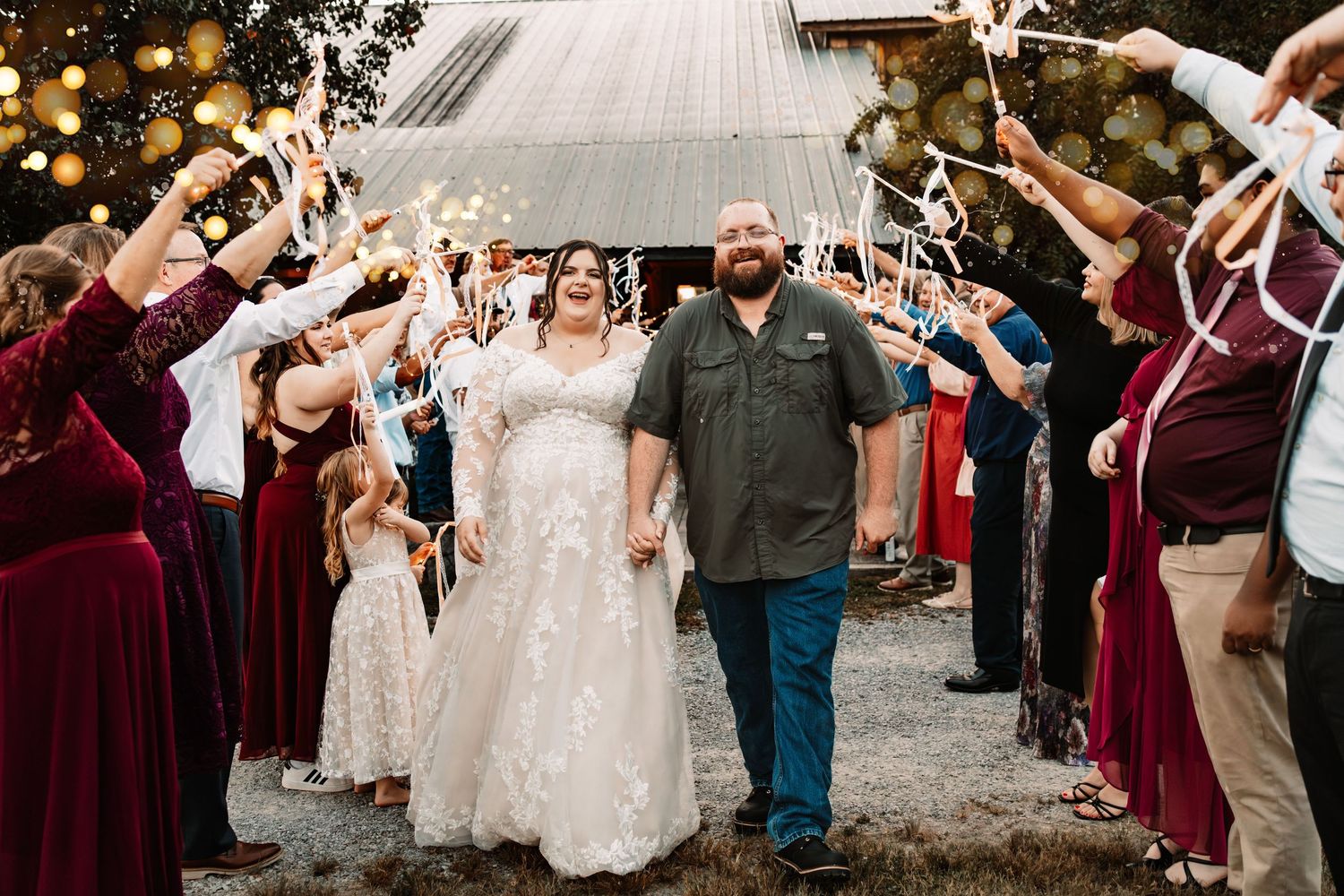 Bride and groom walk through sparkler send-off tunnel with wedding guests at rustic barn reception.