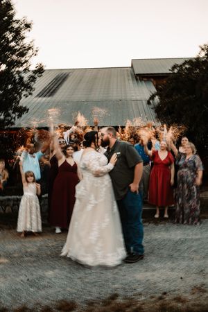 Bride and groom kiss surrounded by guests holding sparklers outside a rustic barn at sunset wedding.