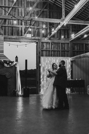 Black and white photo of bride and groom sharing first dance in a rustic barn with string lights overhead.