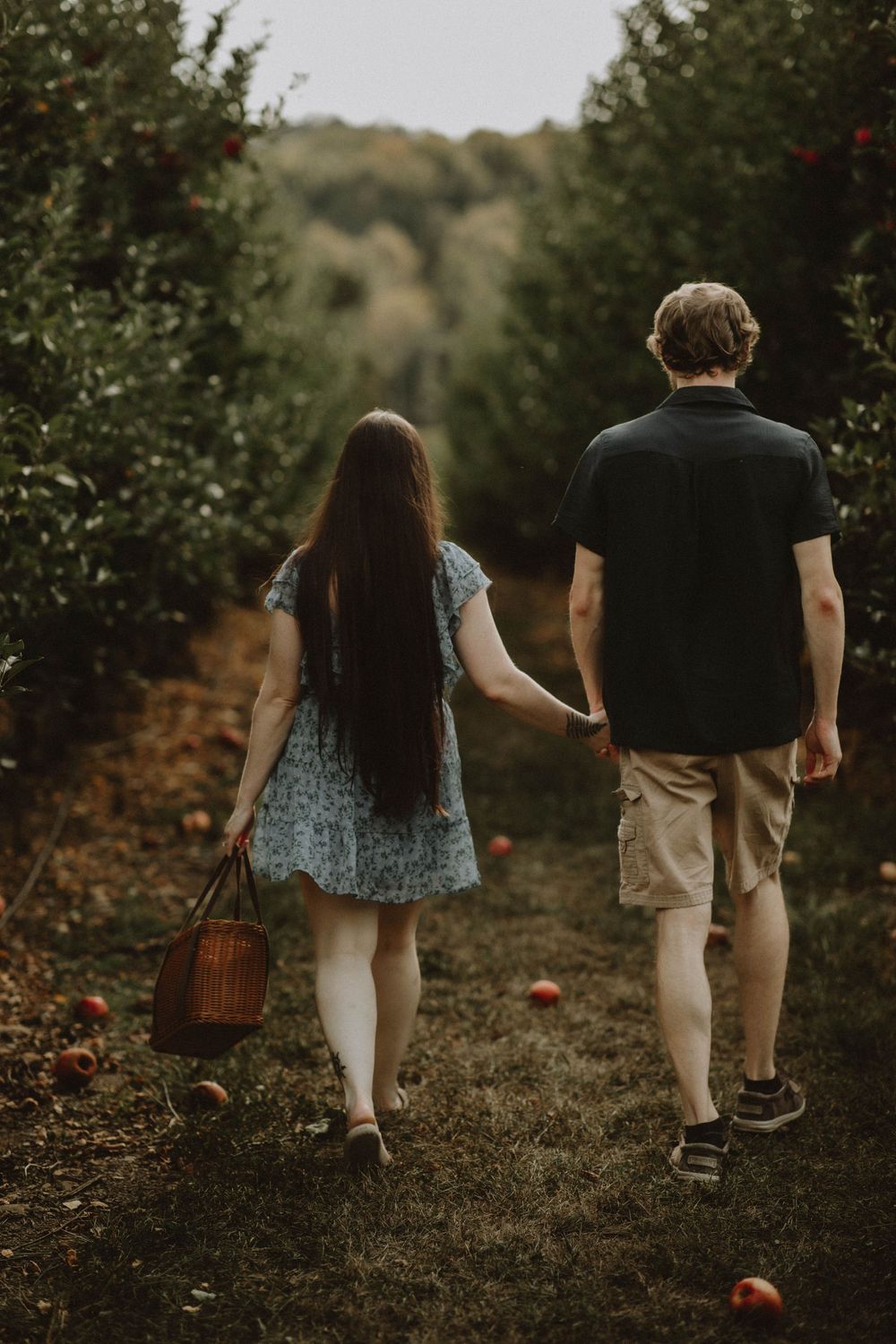 A couple walks hand in hand along a wooded trail carrying a woven basket during autumn season.