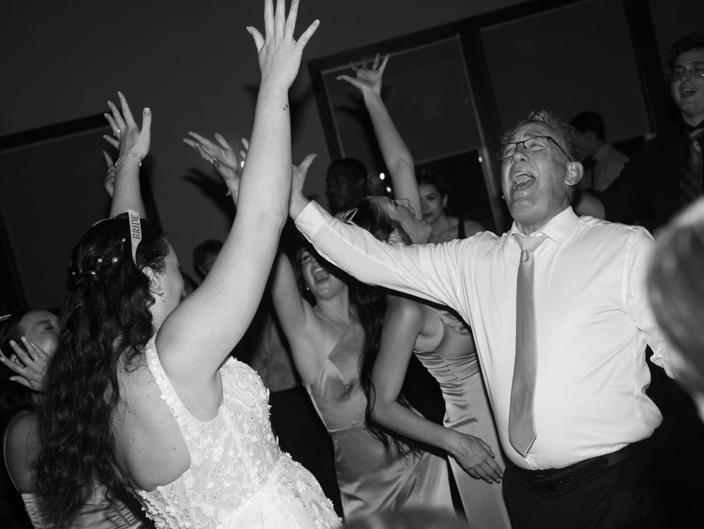 Black and white photo of bride and groom dancing joyfully with guests at a lively wedding reception.