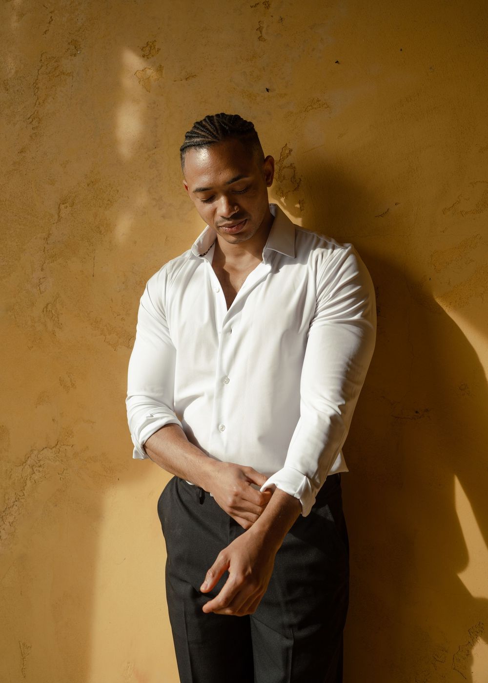 Man in white dress shirt and black pants leaning against a textured yellow wall, looking down thoughtfully.