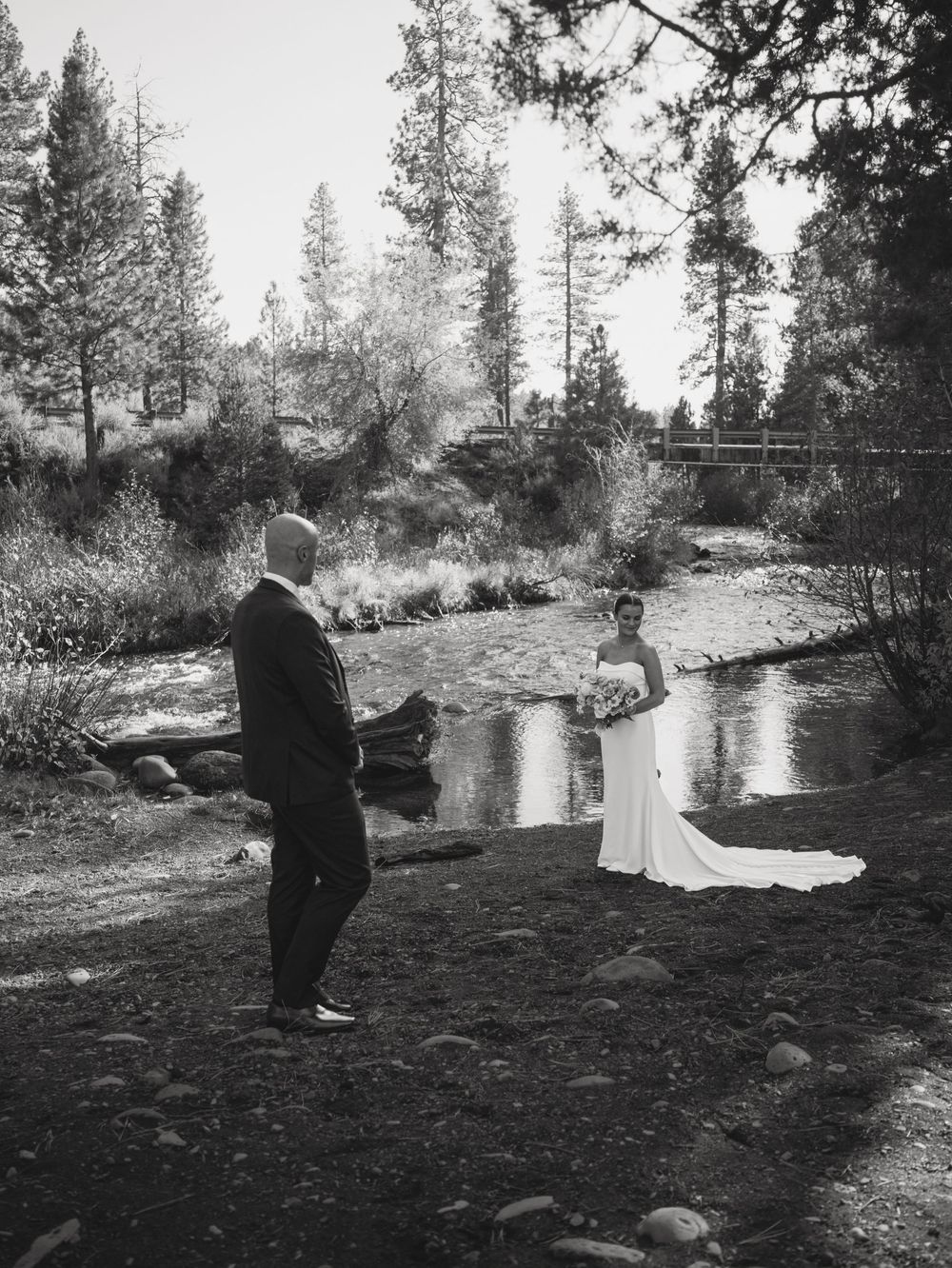 Black and white photo of a bride and groom sharing a first look moment by a serene creek surrounded by tall pine trees.
