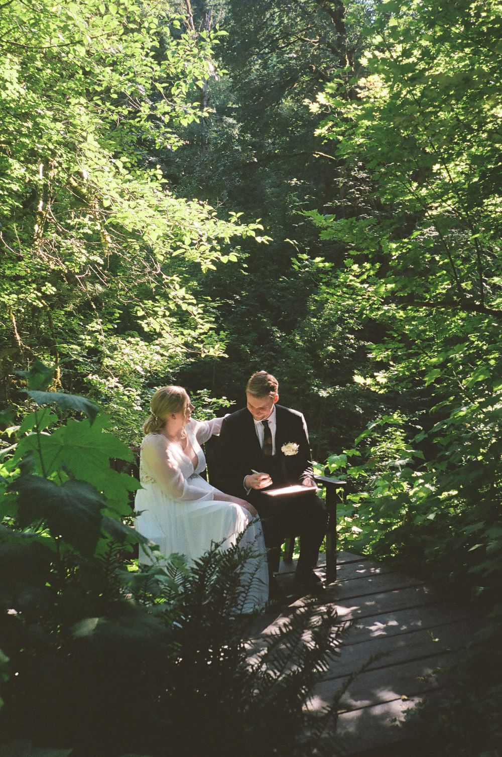 A couple and their wedding party walk along a shaded woodland path during an outdoor ceremony.
