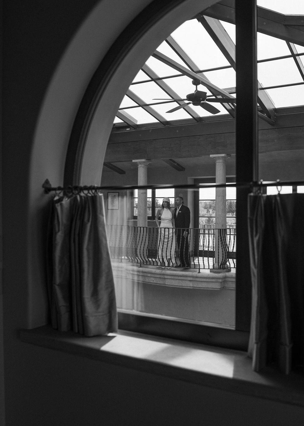 Black and white photo of a couple standing on a balcony viewed through an arched window with curtains.
