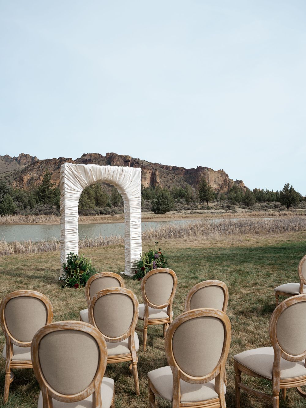 Outdoor wedding ceremony setup with elegant Louis XVI chairs facing a white draped arch, set against a scenic rocky landscape.