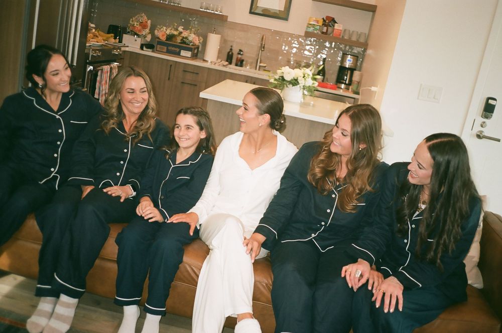 Bride in white robe sits with bridesmaids wearing matching dark navy pajamas, laughing together indoors.