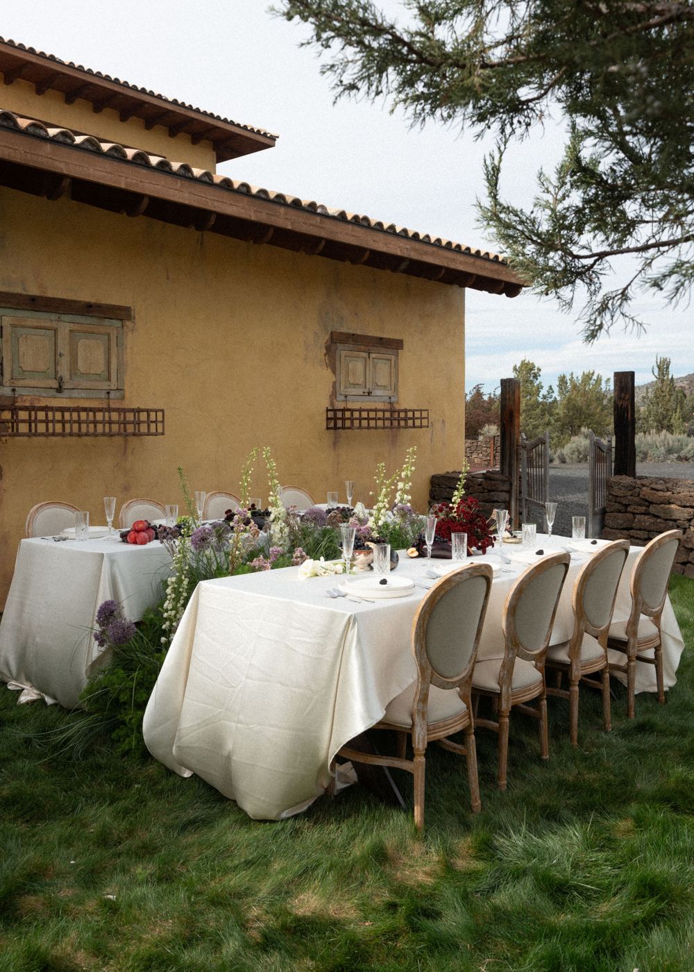Elegant outdoor dining table with white linens and floral centerpieces set on green grass beside a rustic adobe building.