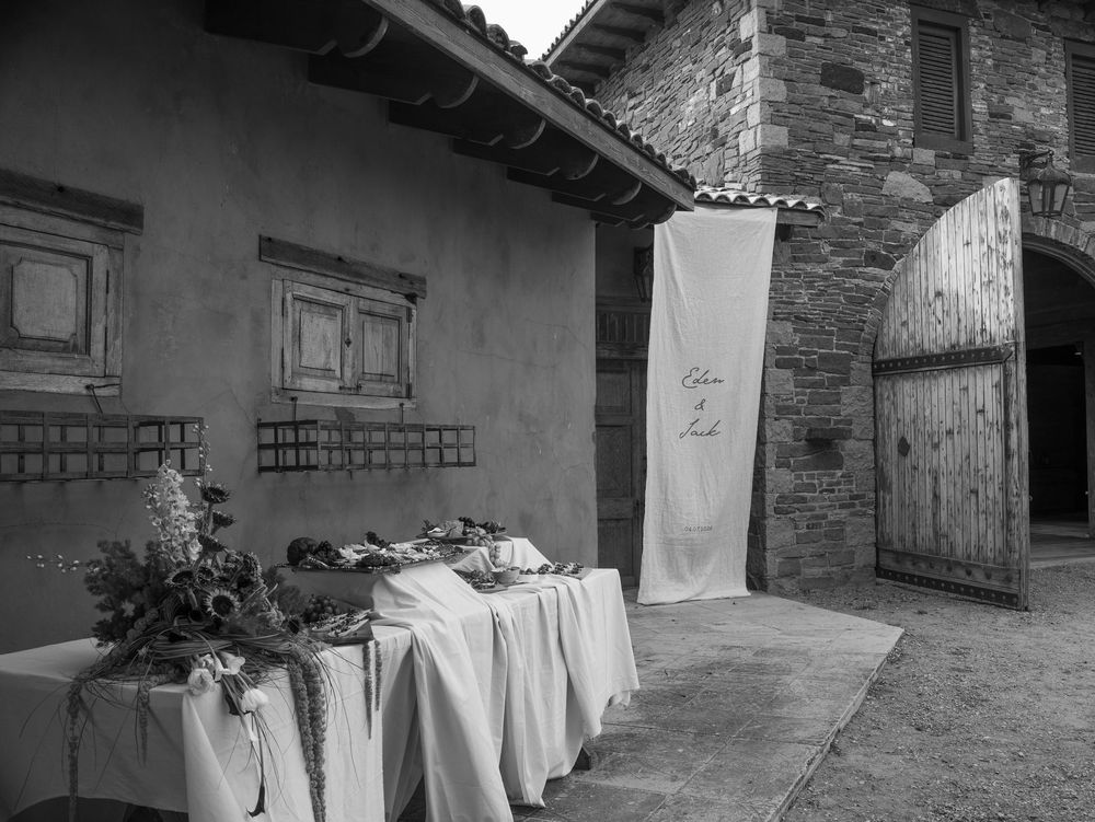 Black and white photo of an outdoor rustic wedding reception table with floral arrangements near a stone building.
