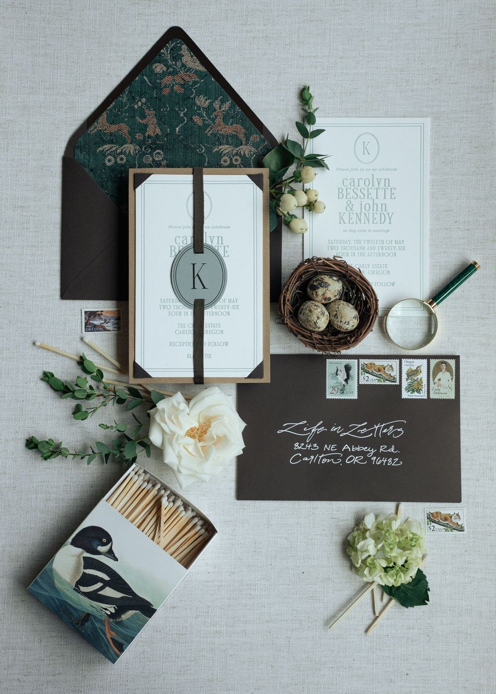 Elegant wedding invitation suite with dark envelope, monogram, bird art matchbox, quail eggs, and floral accents on linen background.