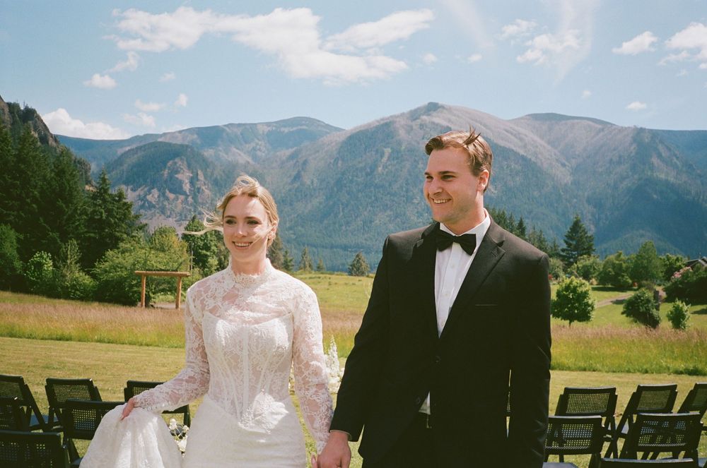 A bride in lace gown and groom in tuxedo stand before stunning mountain backdrop at outdoor wedding ceremony.