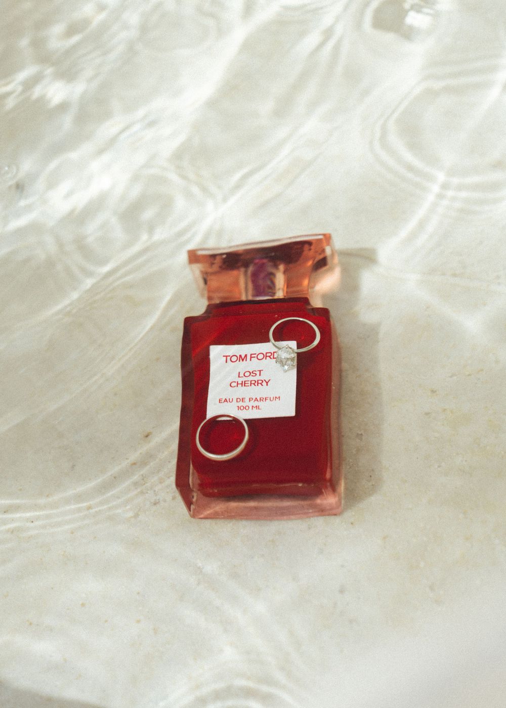 Tom Ford Lost Cherry Eau de Parfum 100ml red bottle with rings resting on it, placed on a reflective wet surface.
