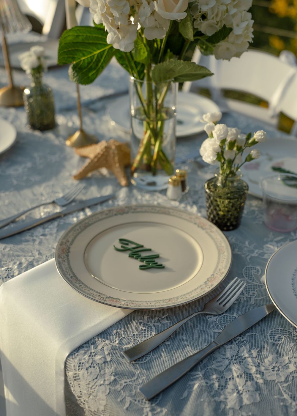 Elegant wedding table setting with lace tablecloth, white floral centerpiece, fine china plate with green name card, and starfish decor.
