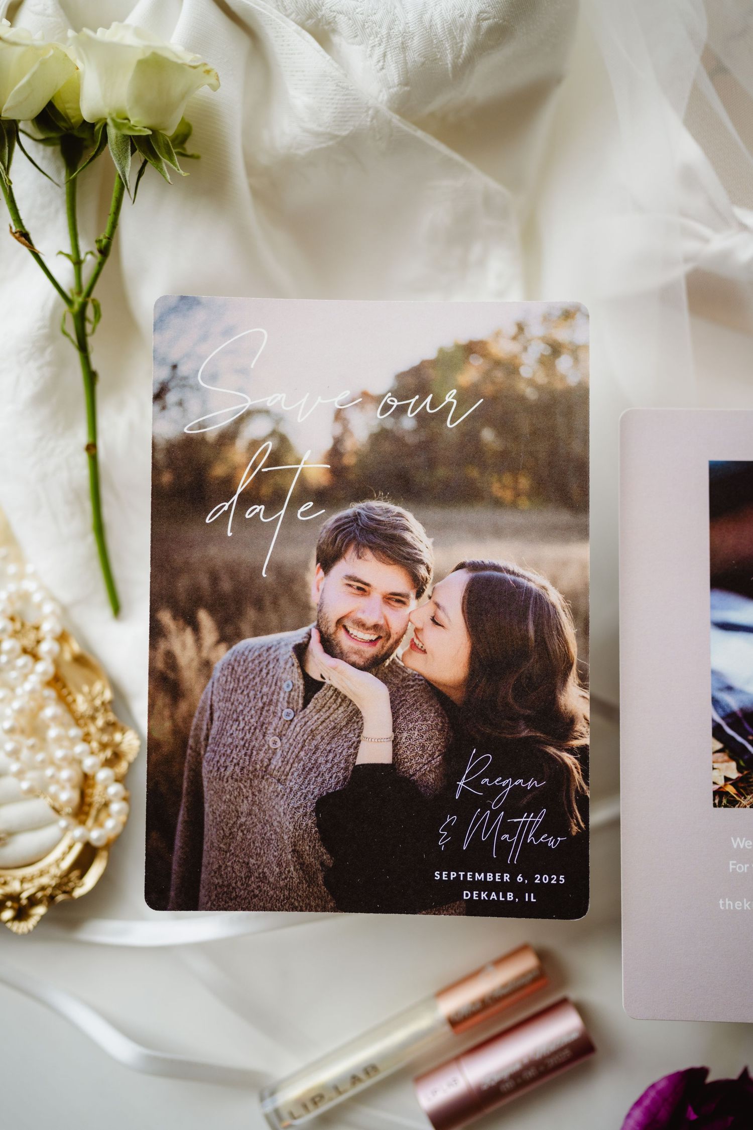 Save the date cards and romantic photography elements laid out on white linen with flowers and cosmetic products.