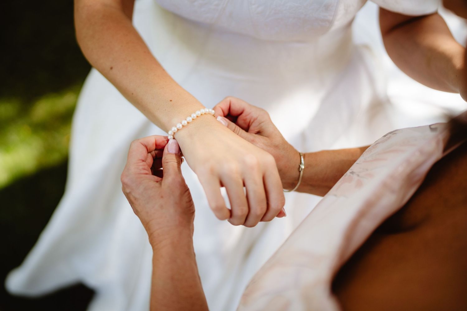 Close-up shot of hands adjusting a delicate silver bracelet against a white dress in natural outdoor lighting.