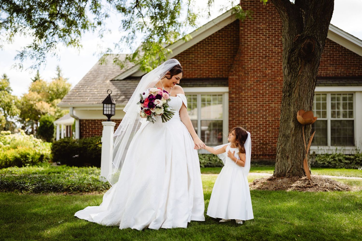 Two people dressed in elegant white gowns stand on a lush green lawn in front of a brick home.