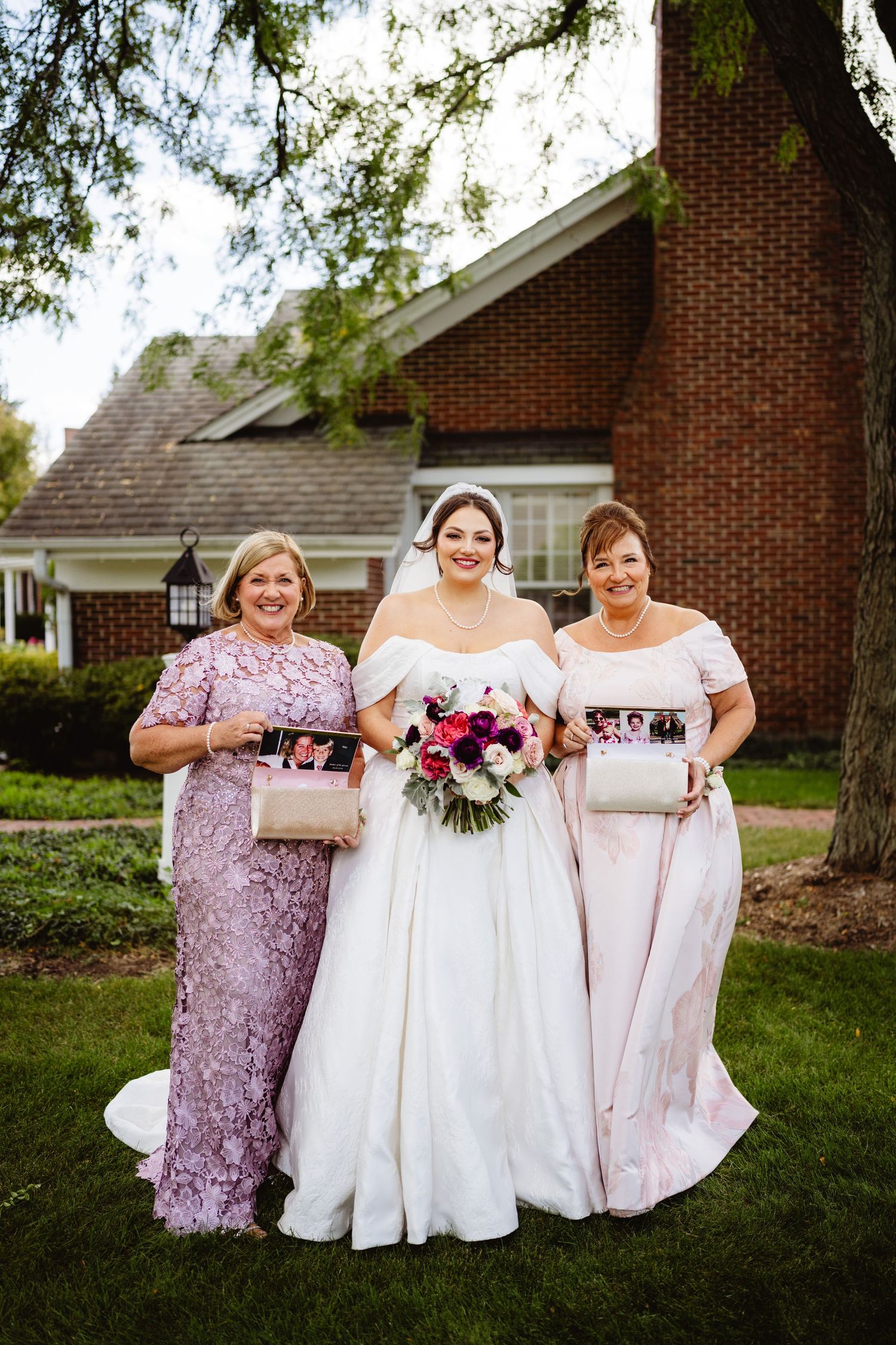 Wedding party members in pink and white dresses pose together on green lawn by brick church building.