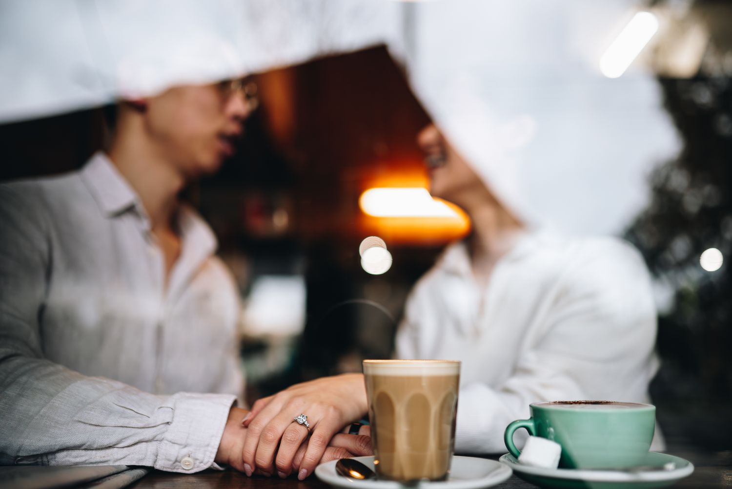 Two people share coffee and conversation at a cafe table holding hands and showing off her engagement ring during a Melbourne photoshoot