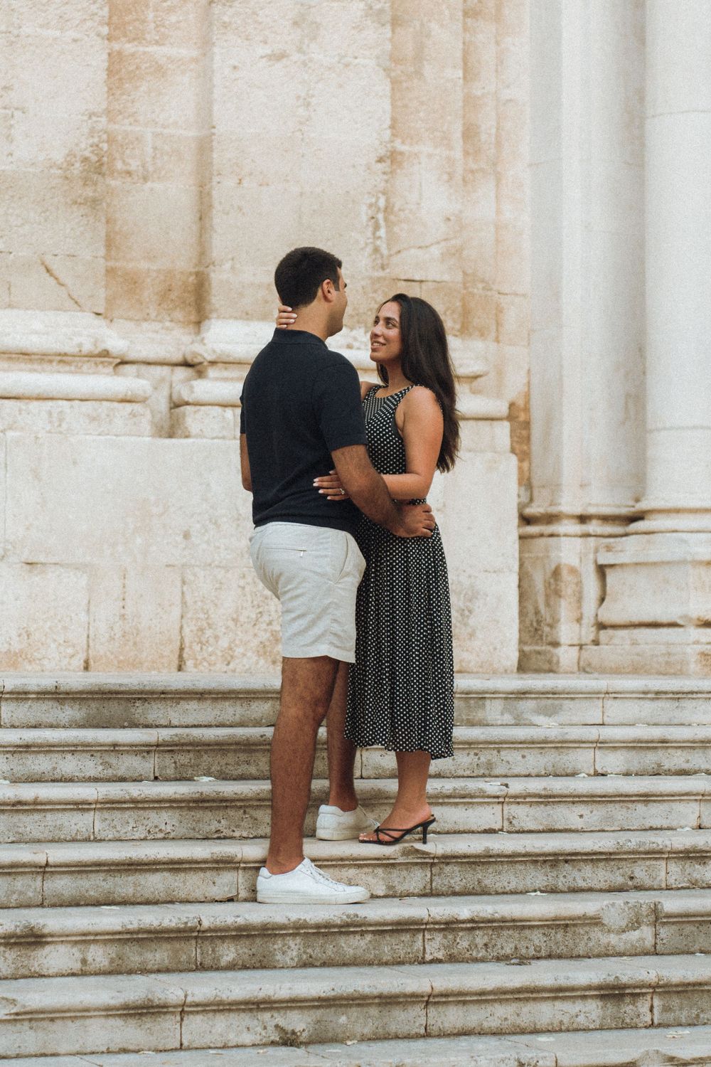A romantic couple embraces on marble steps in front of a classic stone building architecture.