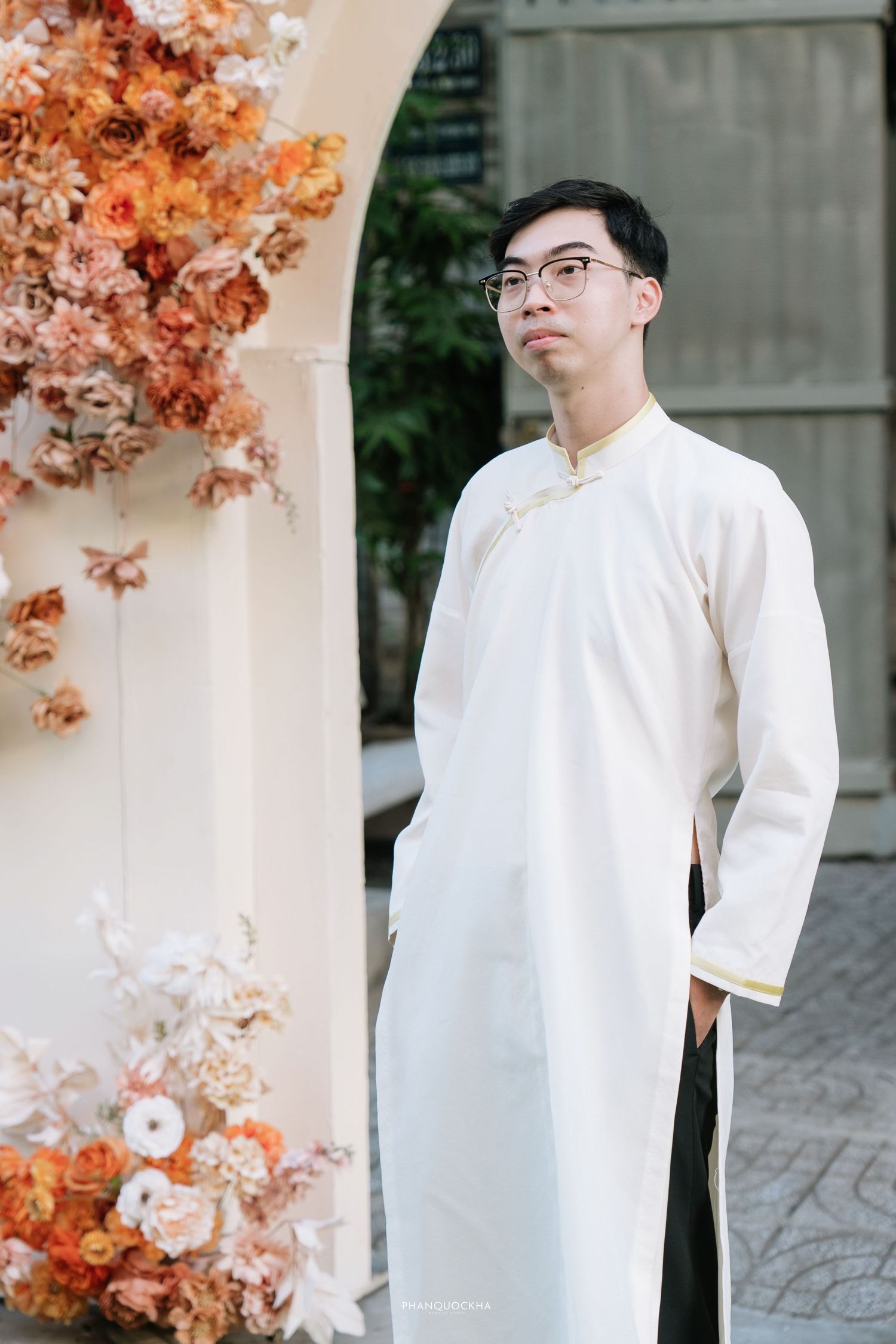 Young Vietnamese man in white ao dai posing beside orange floral arch at wedding ceremony entrance.