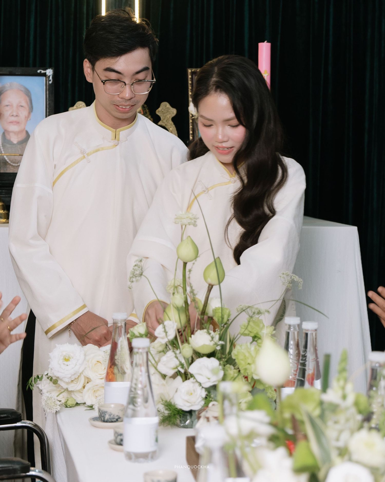 Young couple in white traditional Vietnamese ao dai arranging flowers at a ceremonial altar table.