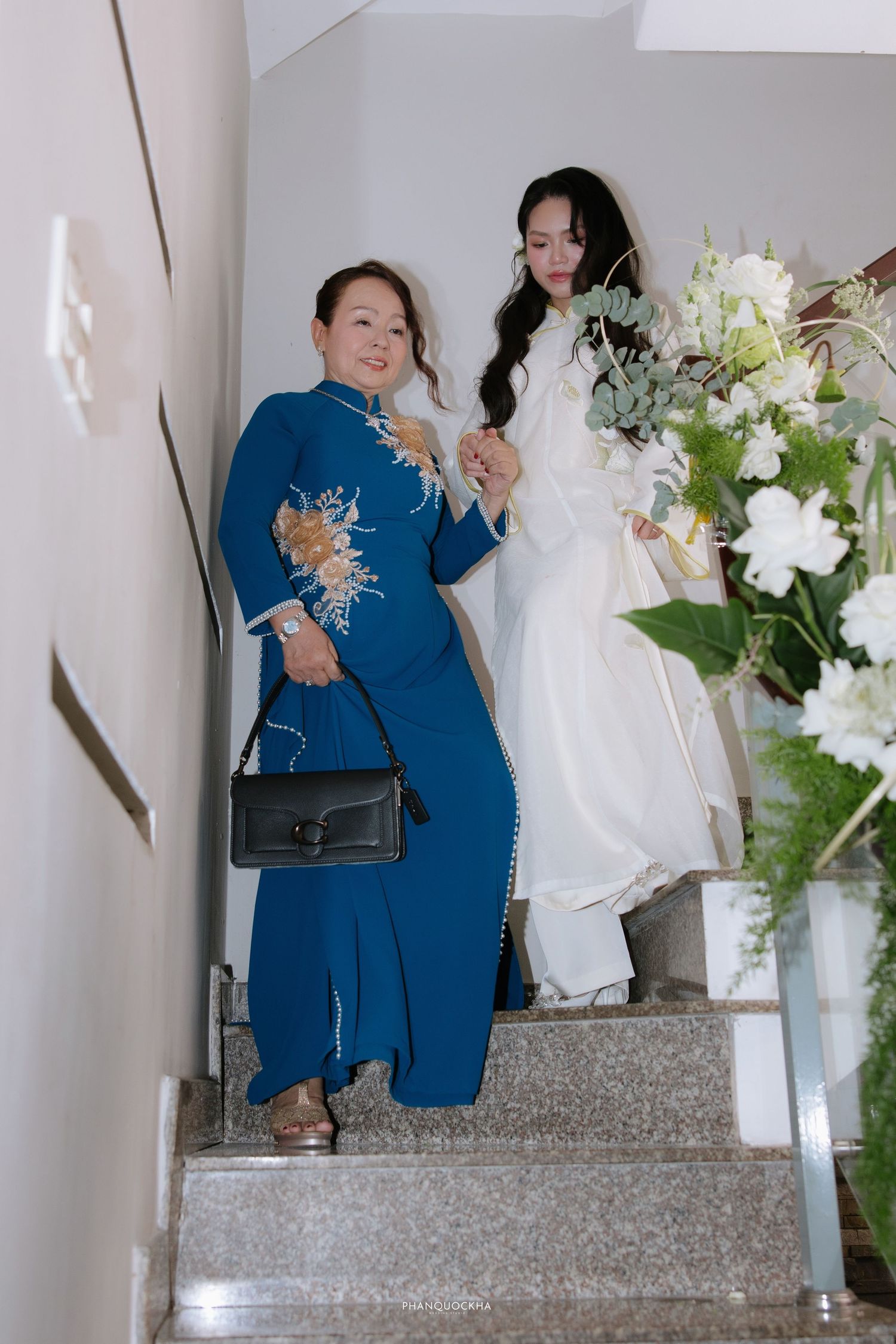 Two women in Vietnamese ao dai descend stairs, one in blue with floral embroidery, one in white bridal gown holding bouquet.