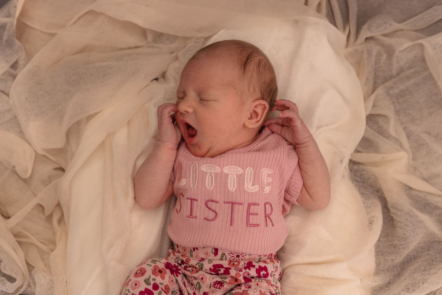 Sleeping newborn baby wearing pink 'Little Sister' onesie on cream colored fabric.
