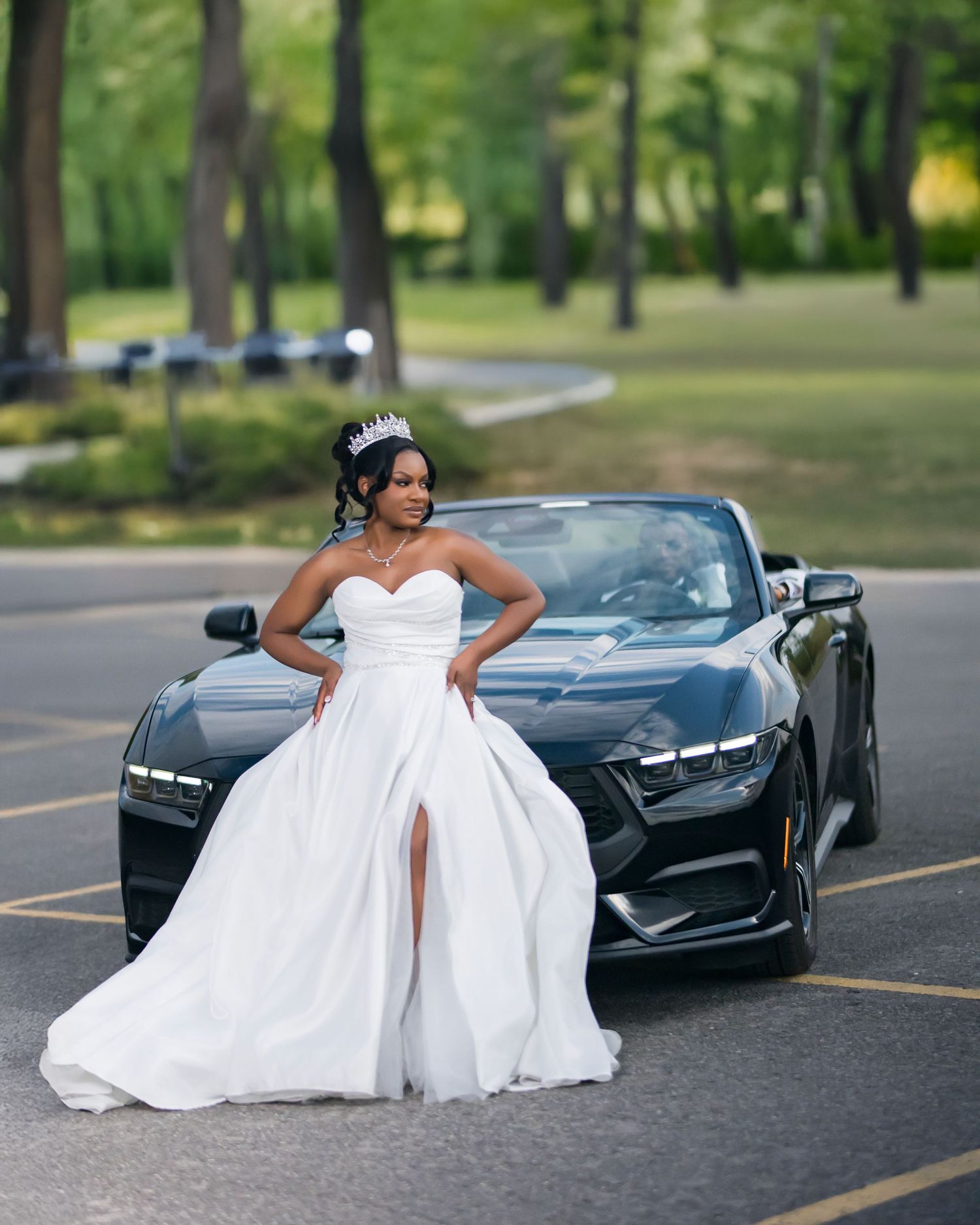 Someone in a white bridal gown poses elegantly next to a sleek black sports car in a sunny outdoor setting.