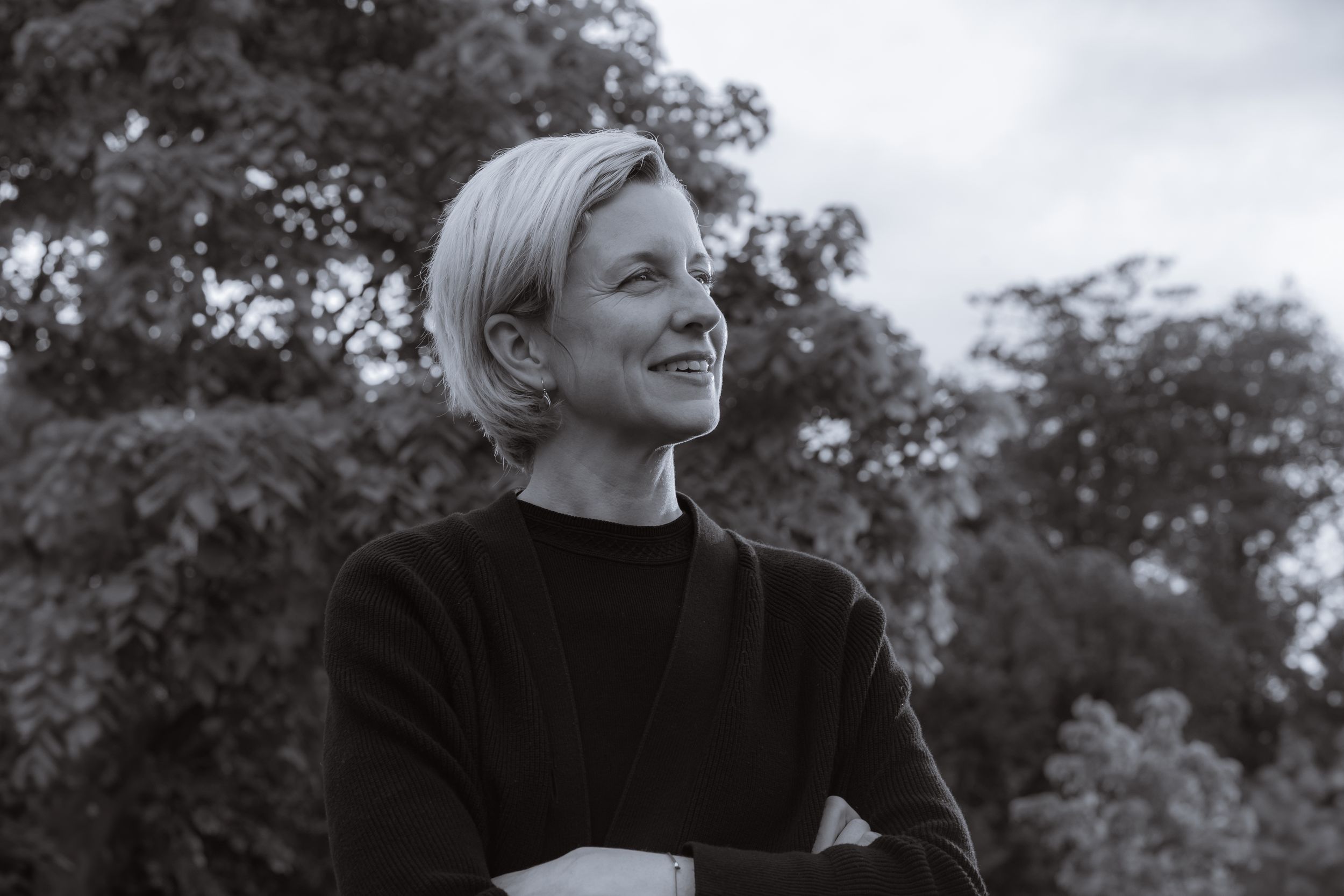 A lady looking to the right, smiling, in black and white with trees and sky in the background