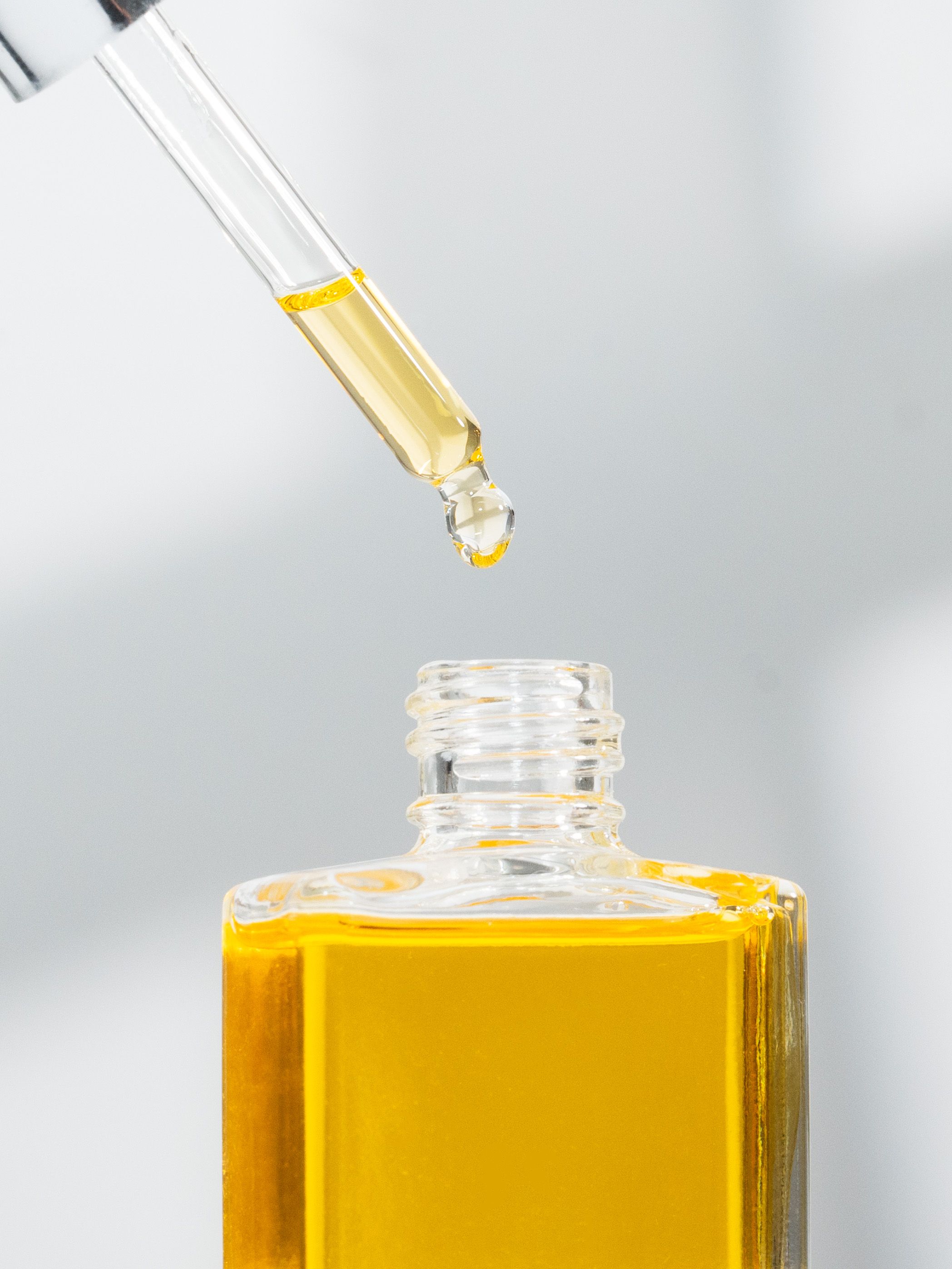 Golden liquid oil drops falling from a glass dropper into a small glass bottle against a light gray background.
