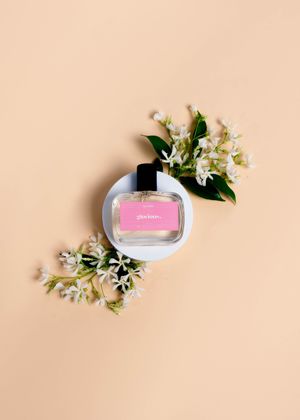 Perfume bottle with pink liquid surrounded by delicate white flowers and green leaves on peach background.
