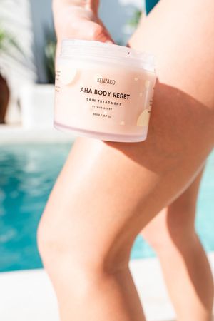 Close-up product shot of pink AHA Body Reset skincare bottles against a blurred turquoise pool background.