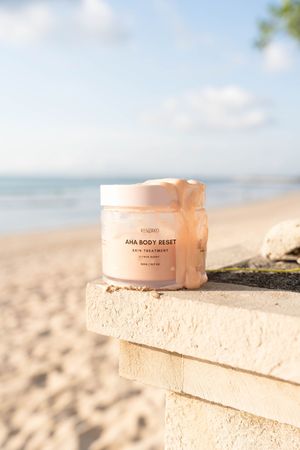 Pink skincare product containers arranged on sandy textured stone blocks at a beach with ocean in the background.