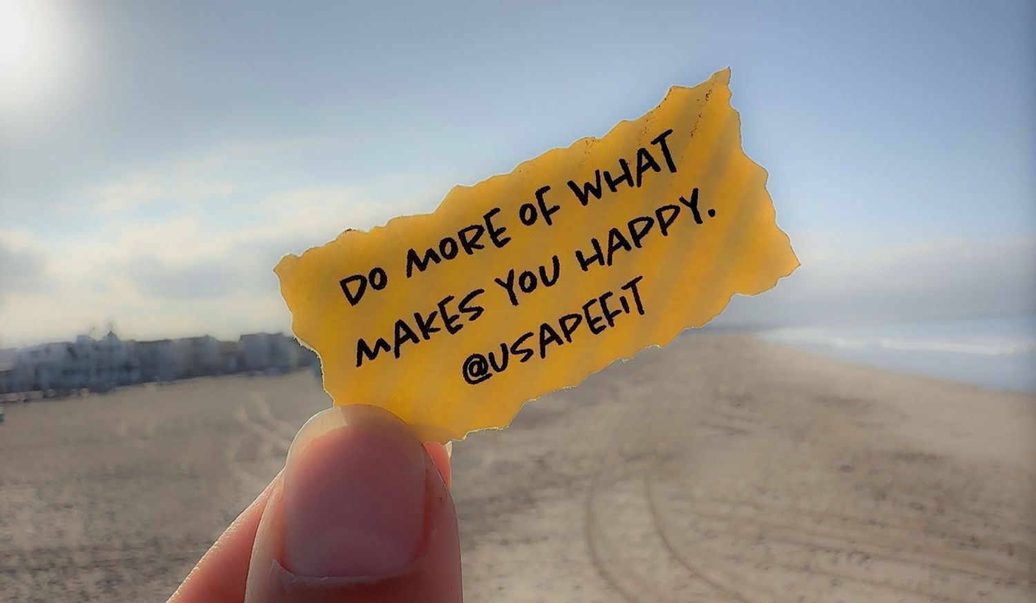 Small torn paper note with inspirational handwritten message is held up against blurry sea and sky background.