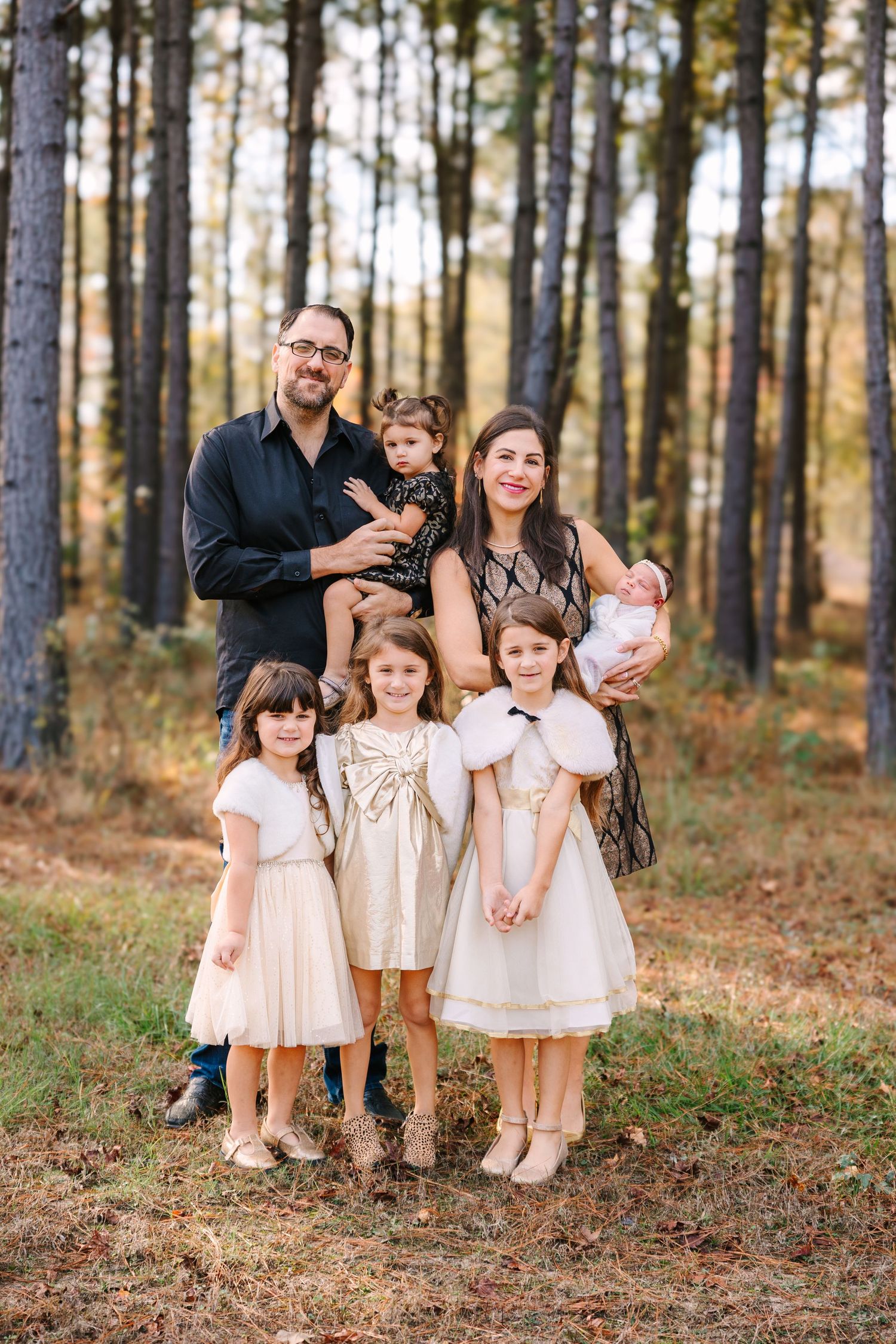 A group wearing light-colored dresses poses together in a sunlit pine forest during autumn.