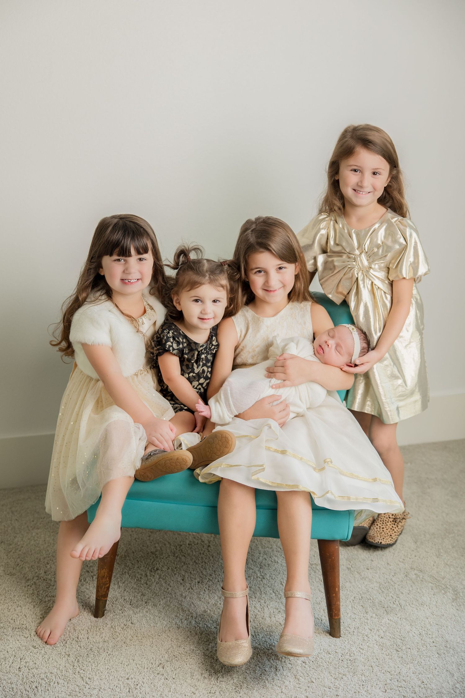 Children wearing special occasion dresses gather on a turquoise chair while holding a baby in a formal portrait setting.