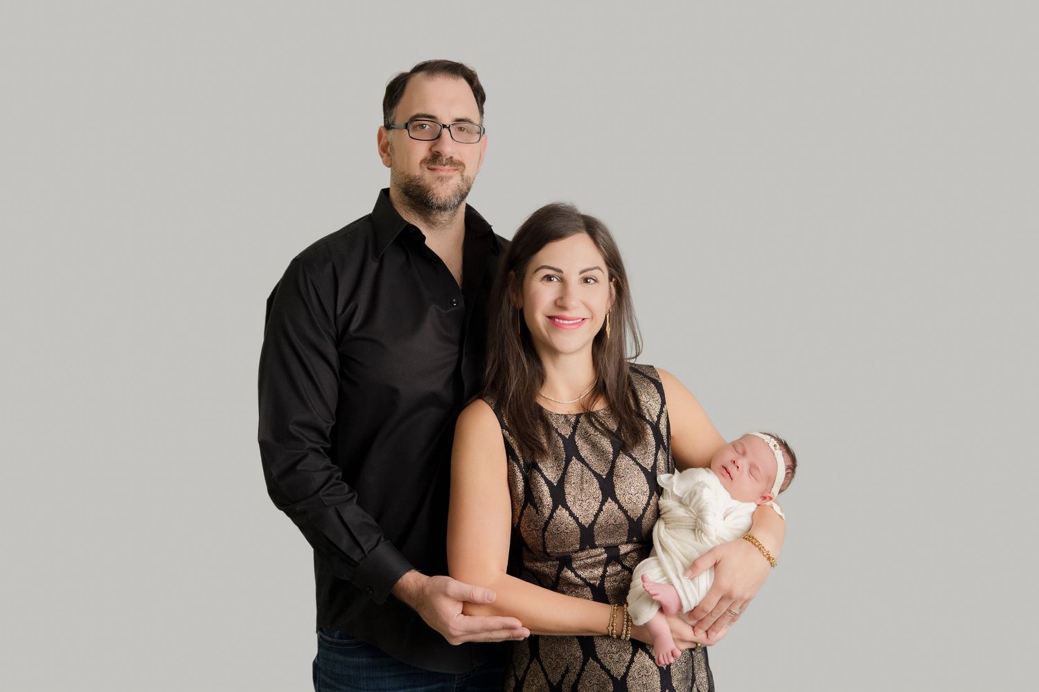 A family portrait session captured in studio with parents cradling their newborn baby against a grey backdrop.